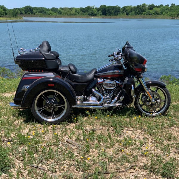 Black trike motorcycle parked on a grassy area near a body of water with trees in the background.