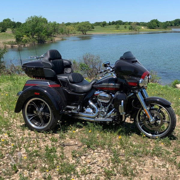 Black trike motorcycle parked on a grassy area near a body of water with trees in the background.